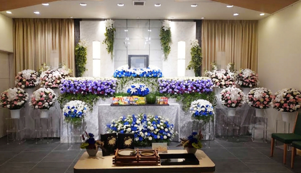 青と白のユリや菊を中心に彩られた生花祭壇。手前には焼香台と供物が配置された式場の様子。