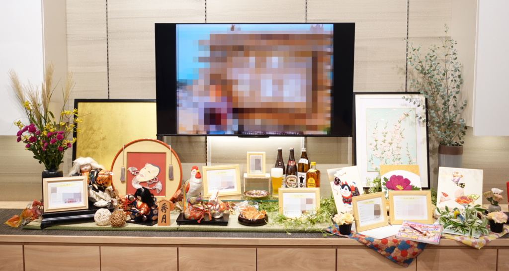 葬儀会場のメモリアルコーナー。中央の大型モニターと周囲に飾られた伝統的な人形、写真立て、好きだったお酒やビール、絵画作品、季節のお花などの思い出の品々